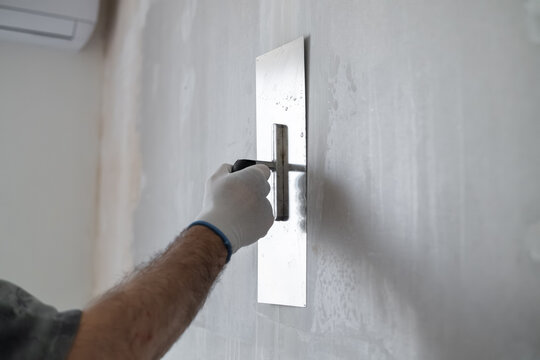 Finishing contractor using stainless steel trowel to apply a smooth layer of plaster on interior wall. Clean workspace with bright lighting and minimal distractions visible in background.