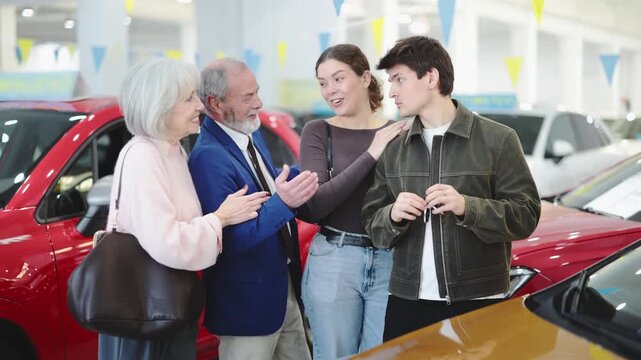  Elderly man and woman hand over car keys to young couple during gift or purchase of car from car dealership