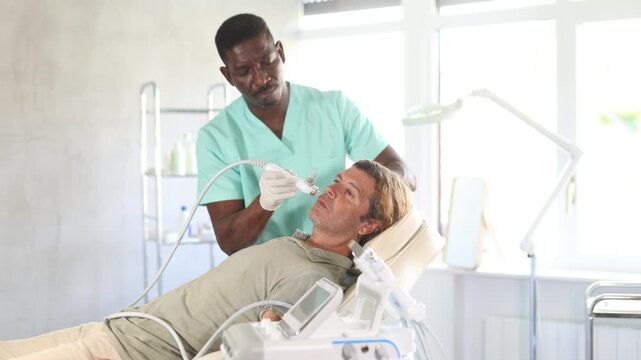 During visit to cosmetology office, client man undergoes facial skin moisturizing procedure using water-oxygen sprayer to smooth dermis and saturate deep layers of skin with moisture