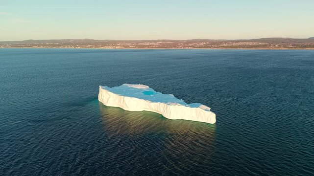 Aerial drone view of a medium tabular iceberg with a circular turquoise melt pool glowing in warm evening light floating off the Newfoundland coastline