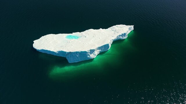 High aerial drone view looking straight down at a large tabular iceberg with a turquoise melt pool drifting in dark deep ocean off Newfoundland