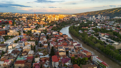 Obraz premium Breathtaking aerial sunset over Tbilisi, Georgia, showcasing the urban sprawl and red rooftops of the old city districts bathed in intense golden hour light with mountain silhouettes