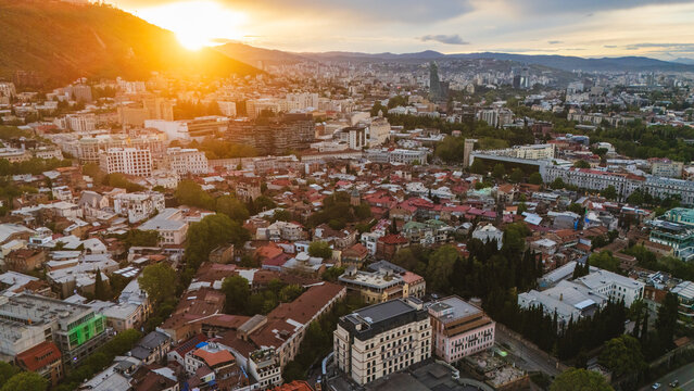 Breathtaking aerial sunset over Tbilisi, Georgia, showcasing the urban sprawl and red rooftops of the old city districts bathed in intense golden hour light with mountain silhouettes