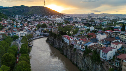 Aerial sunset view of the Metekhi district in Tbilisi, Georgia, with historic houses perched on high cliffs above the Kura River under a warm golden glow © Michele