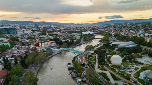 Aerial view of Rike Park in Tbilisi, Georgia, featuring the futuristic Concert Hall tubes, the Holy Trinity Cathedral on the hill, and the urban skyline at dusk over the Kura River.