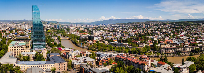 Wide aerial panorama of the Tbilisi skyline in Georgia, featuring the modern glass Biltmore Hotel skyscraper, the winding Kura River, and the sprawling urban landscape under a bright blue sky © Michele