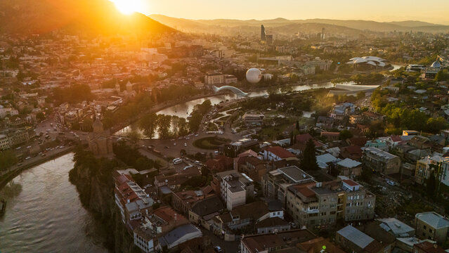 Aerial sunset view of Metekhi district in Tbilisi, Georgia, featuring historic houses on the Kura River cliffs with warm golden hour light over the city