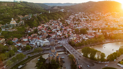 Golden sunset over Tbilisi, Georgia, featuring a wide aerial view of the Kura River, Metekhi Church on the cliff, Rike Park with its hot air balloon, and the iconic Bridge of Peace in the Old Town © Michele