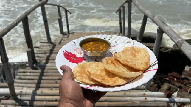 A man holding a traditional breakfast plate of fluffy dal puri and spicy potato sabji, set against a beautiful, sun-drenched seashore background in coastal India.