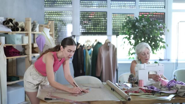 Mature woman creates wardrobe items, sews dresses and suits for clients, makes sweater out of knitwear. Female assistant drawing paper dress pattern by curves