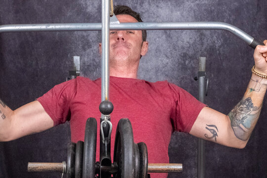 A man performs a back muscle exercise using a multifunction bench. The man is forty-two years old, of Caucasian origin, and wears a burgundy T-shirt