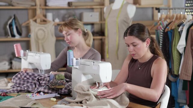 Two female tailors are sitting at a table in the workshop and sewing clothes on machines. Employees of the sewing industry make wear according to individual measurements. High quality 4k footage