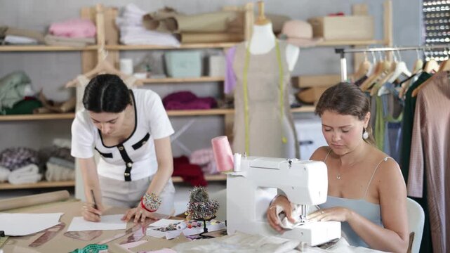 Female dressmakers work together to create custom clothing. Fashion designer stands near a table and draws a sketch on paper next to a seamstress who works on a typewriter. High quality 4k footage