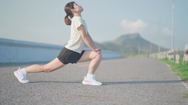 Asian woman with a towel around her neck cooling down and stretching after jogging in the park. Female runner relaxing after morning workout. Fitness, recovery and healthy lifestyle concept