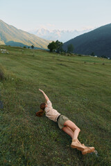 Obraz premium Young woman relaxing on a hillside meadow with mountains in the background, stretching her arm, casual outdoor lifestyle scene suitable for stock, bright atmosphere, natural light