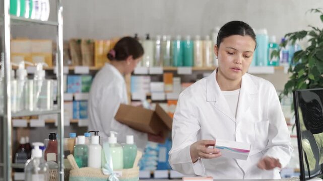 Attentive middle-aged female pharmacist working with computer in drugstore while other restocking in background . High quality 4k footage