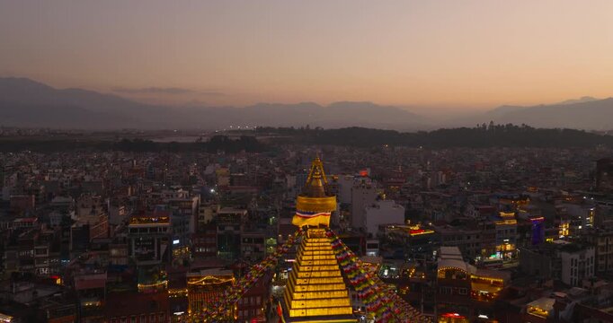Aerial drone reveal starting from the pinnacle of Boudhanath Stupa, a UNESCO World Heritage Site in Kathmandu Nepal, expanding outward glowing stupa, prayer flags surrounding city at fading evening.