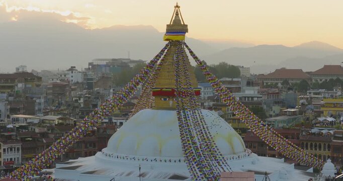 Drone shot of Boudhanath Stupa, a Buddhist monument in Kathmandu Nepal, with prayer flags, sunset sky, city buildings and surrounding hills.