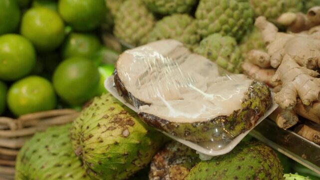 Close-up of sliced soursop on tray wrapped in plastic film surrounded by limes and ginger at a fresh produce market.
