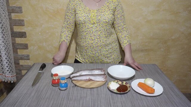 Ingredients for cooking fish dish on the kitchen table. A woman stands near the kitchen table, on which the ingredients for the culinary recipe are laid out. Raw peeled hake fish carcasses lie on a wo