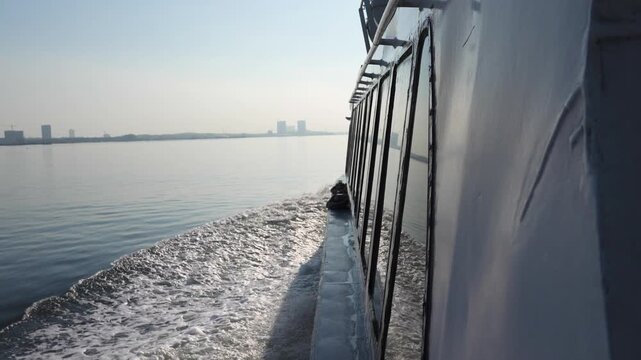 A point-of-view shot from a moving ferry boat deck showing a white foam wake and a distant urban skyline along a major shipping lane in Southeast Asia or Strait of Malacca. Travel and maritime themes.