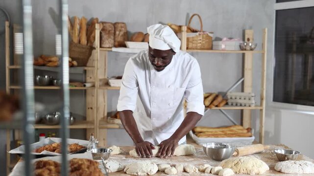 Skilled black baker expertly rolling dough into long loaf shape, preparing for baking in warm rustic bakery environment, surrounded by freshly baked bread and pastries arranged on wooden shelves