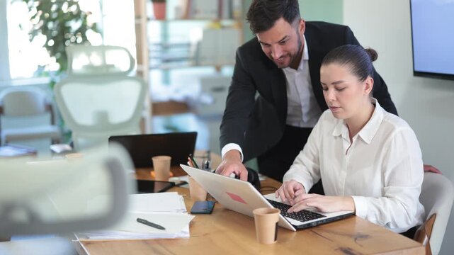 IT specialist man helps a young female employee work on a laptop against the backdrop of the interior of a modern office. Team of web designers is working on a new website. High quality 4k footage
