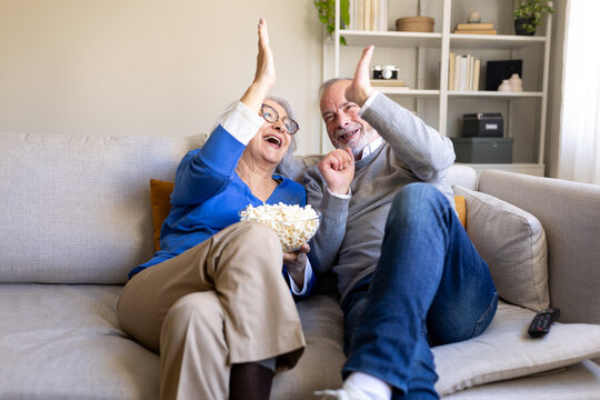 Excited senior couple watching tv and cheering with popcorn on sofa