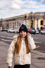 Obraz premium Little Girl Enjoying a Sunny Day While Waiting on a Bustling Street. A cheerful girl stands outdoors on a lively street, wearing a cozy white jacket and a black beanie.
