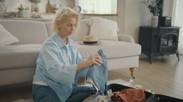 Old woman preparing to business trip or summer travel in vacation, pack suitcase . Portrait of happy senior lady sitting on floor and putting her clothes into travel bag, dream about merry weekend