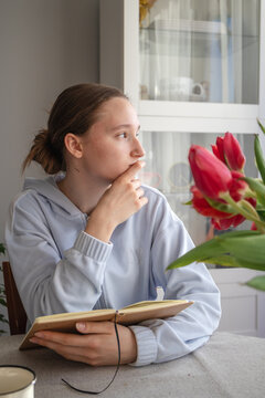 Pensive teenage girl looks away, distracted from reading a book at table near tulips  in warm sunlight. Young student studying at home, preparing lessons and spending quiet educational time indoors.