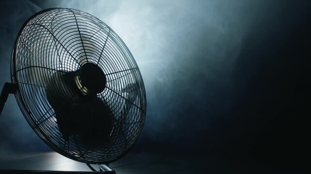 Industrial floor fan in operation against a dark, smoky background with strong backlighting highlighting the moving blades.