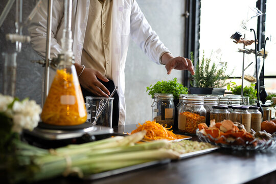 A researcher in a white lab coat performs botanical extraction using distillation glassware surrounded by fresh herbs with Distillation Equipment, ingredients for natural medicine or aromatherapy