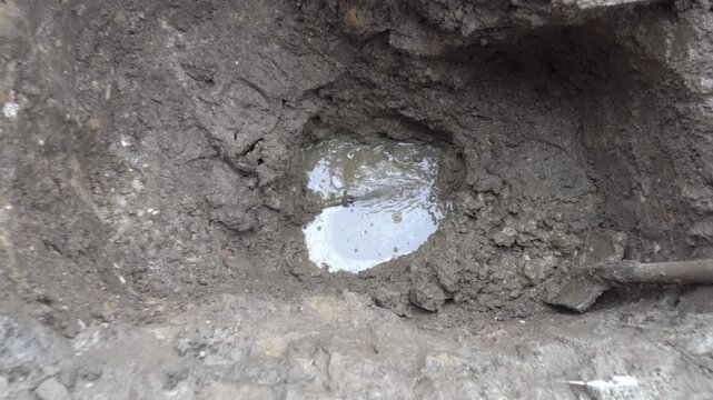 Water main break and repair works in a muddy trench, A deep hole in muddy soil during emergency water pipe repairs. Water sprays under pressure from a ruptured underground pipe, forming a puddle.