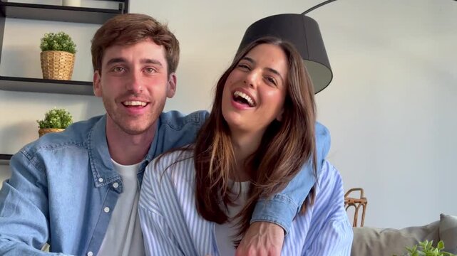 Smiling young couple talking on a video call at home, waving and laughing at the camera while sitting together on the couch in their modern living room in a point of view shot