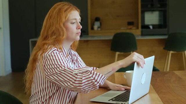 Focused young woman with long red hair wearing pajamas sitting at a wooden table in her kitchen, opening her laptop to start her workday and drink coffee, embracing the remote work lifestyle