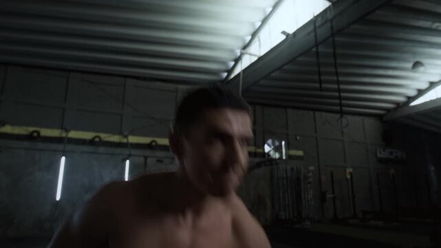 Athletic shirtless man performing kettlebell snatches during intense functional fitness training session inside dim gym hall professional cinematic footage.