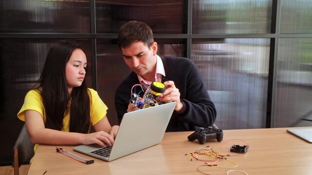 Mentor helps a student build a small wheeled robot while she codes on a laptop; wires and components spread across the table in a modern workspace