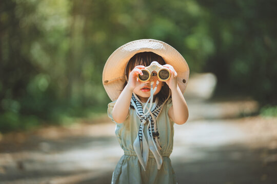 Curious Asian toddler girl looking through binoculars in a sunlit park forest. Little child adventurer exploring nature during summer vacation. Concept of childhood discovery, education, outdoor