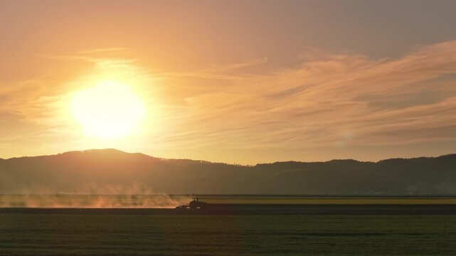 Dynamic tractor tilling field raising backlit dust plume during sunset in Transylvania near Cristian, stunning golden hour landscape with clouds and copy space, static 4K