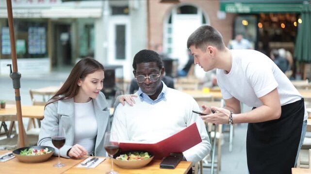 European woman and her African husband are dining on the terrace of a cozy restaurant, they choose food and order from the waiter. International couple on the veranda of a restaurant in a European