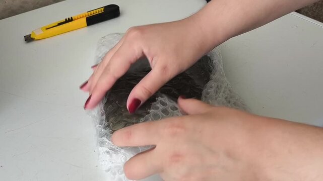 Hands removing bubble wrap from transparent package with pumpkin seeds and white stripe. Clean unboxing and food packaging concept on neutral gray tabletop.