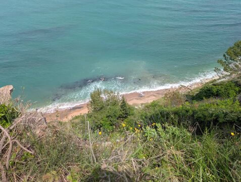 Ripresa dall&rsquo;alto di una costa naturale con vegetazione verde, spiaggia stretta e mare turchese con onde leggere che si infrangono sulla riva