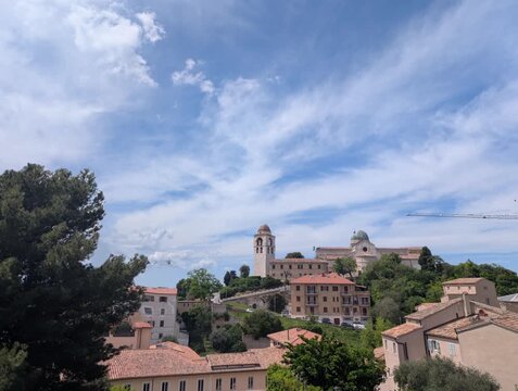  
Veduta luminosa di un borgo marino mediterraneo con tetti in coppi, campanile storico e vegetazione rigogliosa sotto un cielo azzurro con nuvole sparse