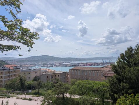 Panoramica luminosa di una citt&agrave; costiera immersa nel verde, con edifici storici, vegetazione rigogliosa e un ampio tratto di mare sullo sfondo sotto un cielo parzialmente nuvoloso