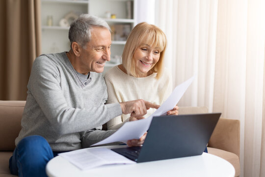 Mature couple checking financial documents with laptop at home, discussing bills, happy senior spouses planning household budget together indoors