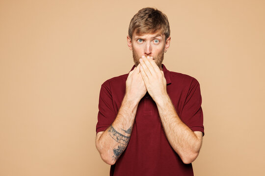 Young sad upset tired irritated shocked man he wearing red shirt casual clothes look camera cover mouth with hands isolated on plain pastel light beige background studio portrait. Lifestyle concept.
