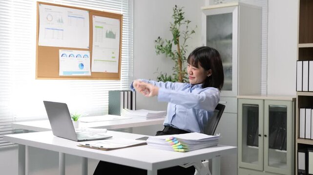 Tired Asian businesswoman stretching her arms and back after working on a laptop computer. Young female office worker relaxing at her desk.