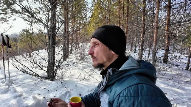 Man In Snowy Forest With Mug Pausing On Pine Trail, Blue Jacket And Black Beanie, Holding Yellow Cup And Small Item, Hiking Poles Planted In Snow Nearby Sunlight Filtering Through Trees,