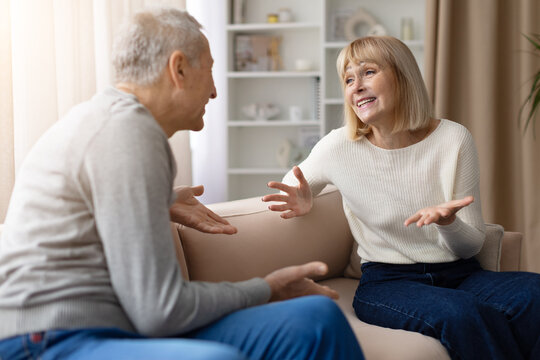 Mature couple sitting on a sofa at home, both gesturing with open hands and talking expressively to each other in a lively conversation in the living room